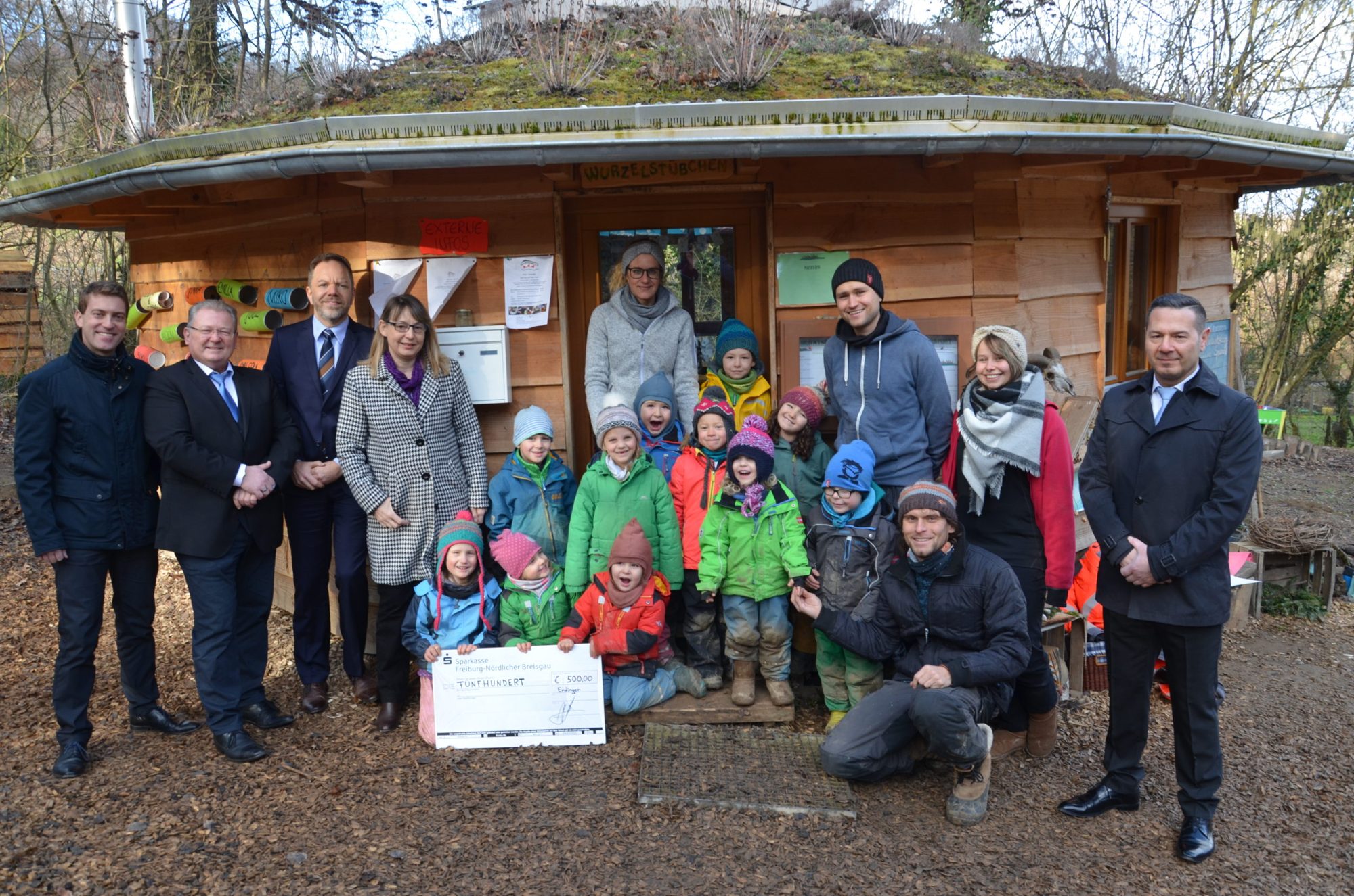 Kinder, Team und Gäste mit Spendenscheck vor dem Bauwagen mit Gründach