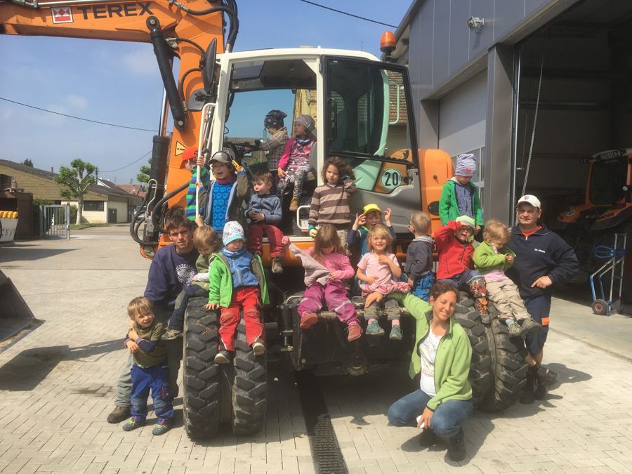 Kindergartengruppe sitzt mit Erzieherinnen und Bauhof-Mitarbeitern auf einem orangen Bagger