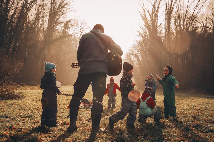 Erzieher spielt Gitarre, Kinder musizieren auf einer Wiese im Wald
